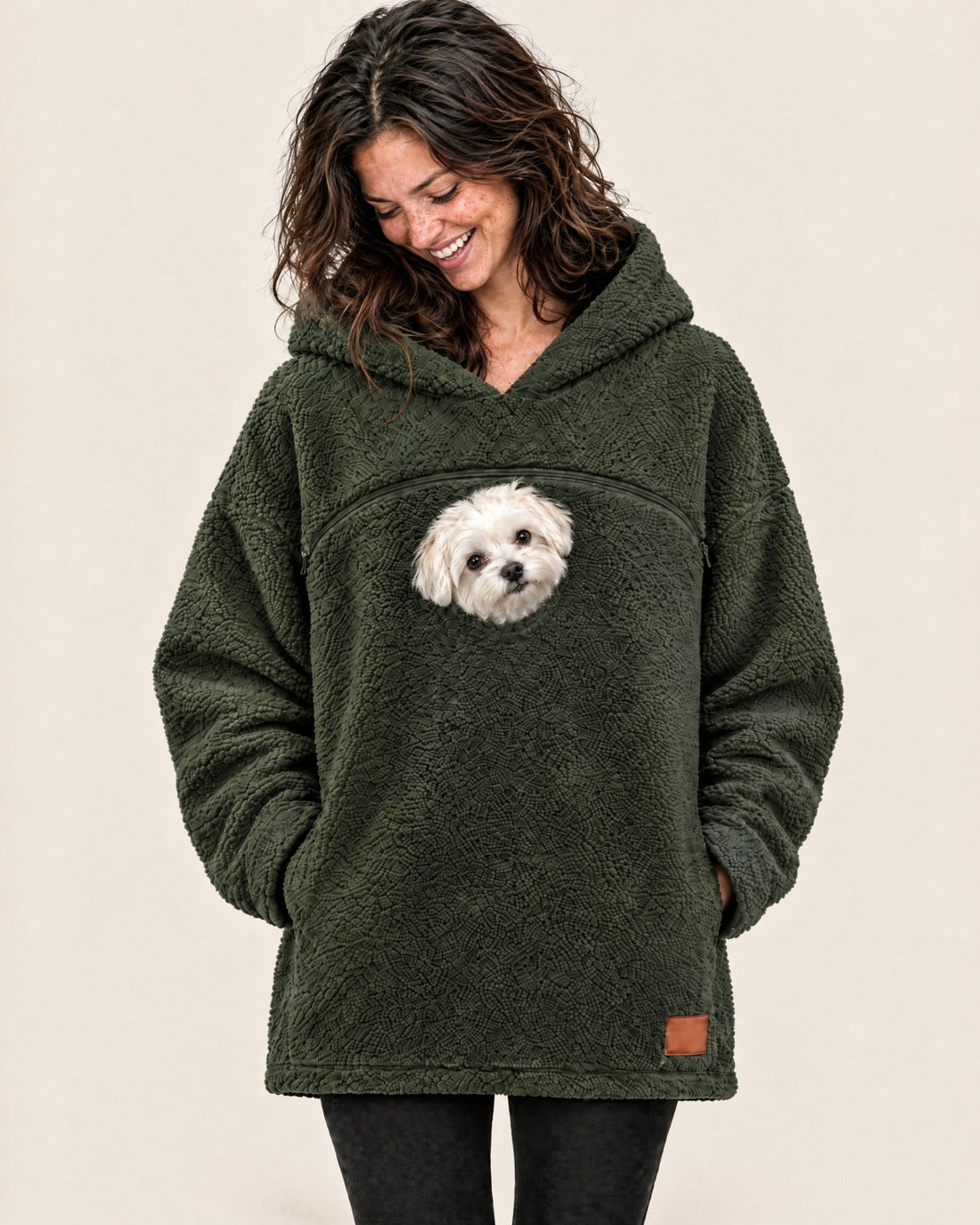 Woman in an army green tucked pouch hoodie laughing as she looks down at the small white Maltese peeking out below the chest zipper