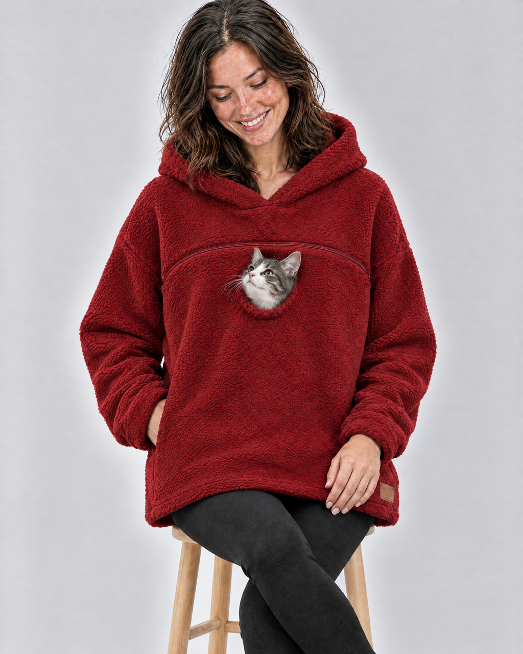 Woman on a stool in a red pouch hoodie smiling down at the grey-and-white kitten peeking out of the chest opening