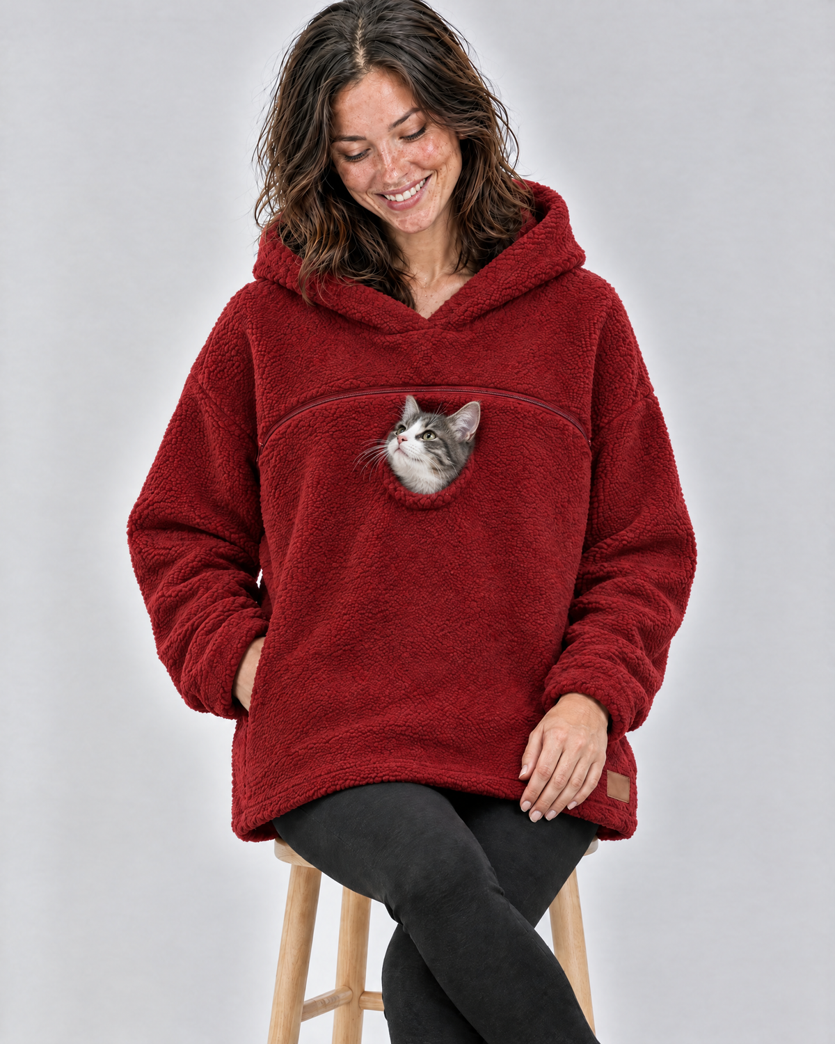 Woman on a stool in a red pouch hoodie smiling down at the grey-and-white kitten peeking out of the chest opening
