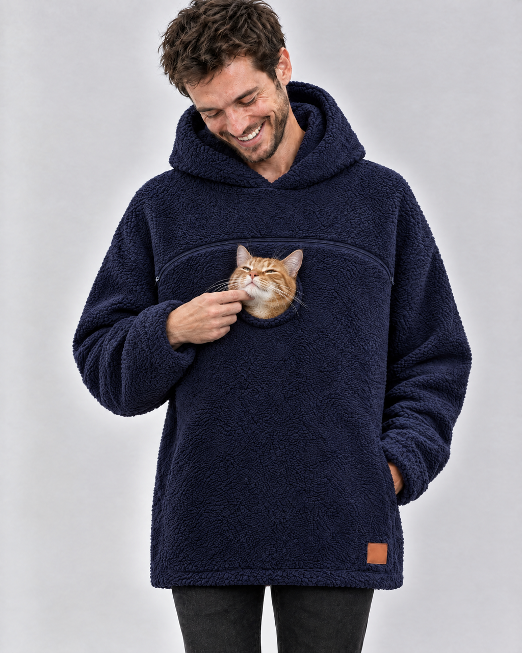 Man laughing in a navy pouch hoodie, scratching the chin of the orange tabby cat peeking out of the chest